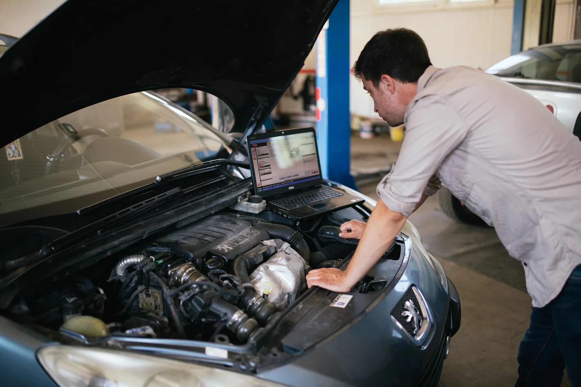 Mechanic using a laptop beside an open vehicle bonnet in a workshop
